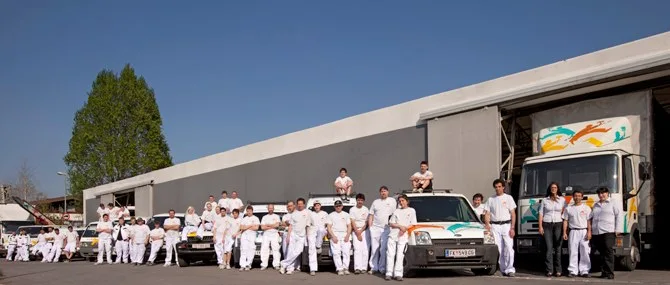 Das gesamte Team der Malerfamilie Petter in weißer Arbeitskleidung vor der Werkhalle und der Fahrzeugflotte