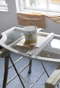 Weißer Fensterflügel auf Böcken, daneben ein Farbkübel und abgedeckter Boden